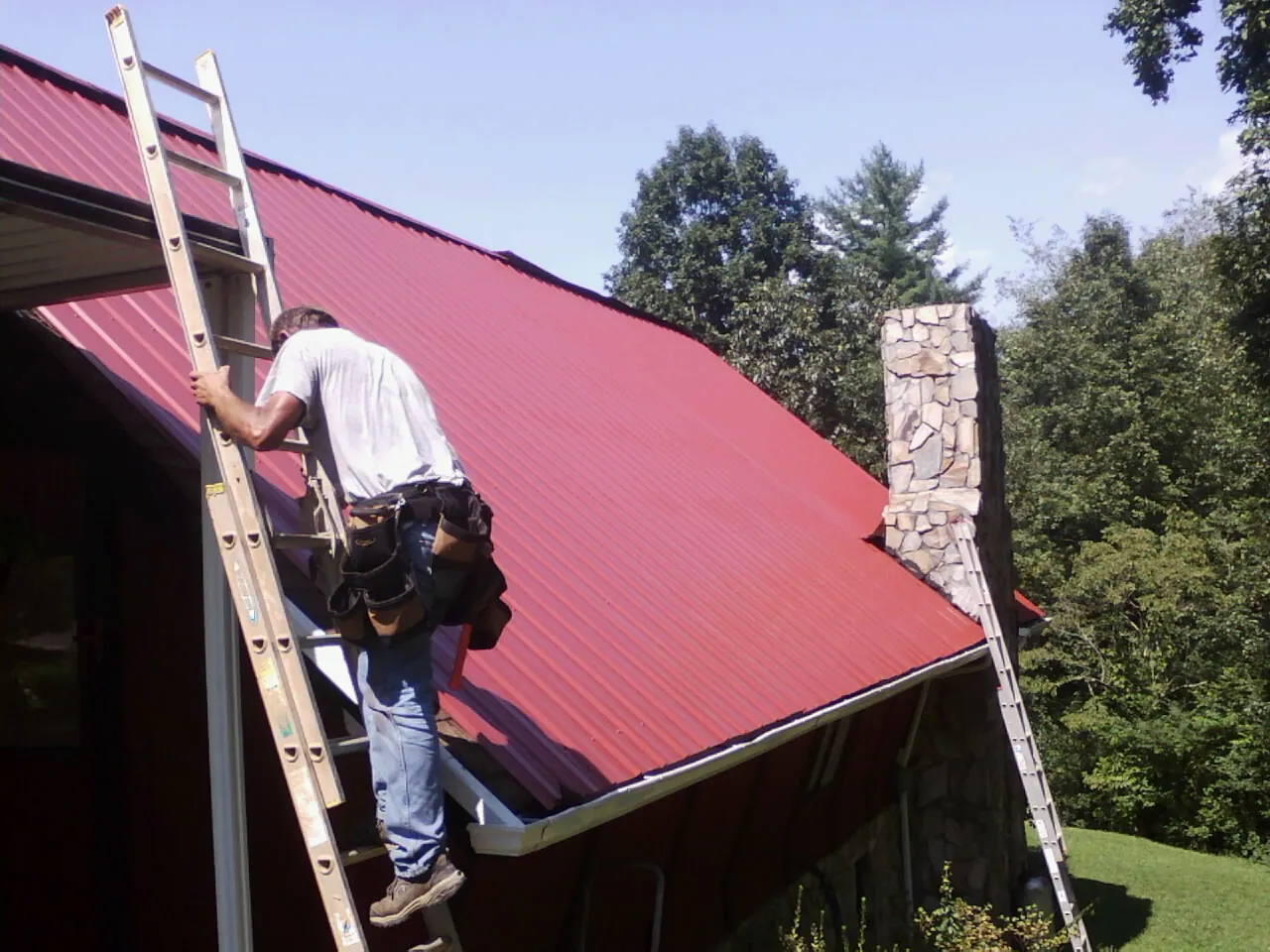Roofer on a ladder beside a red metal roof and stone chimney — Soffit & Fascia Repair by Roofing Brothers in Kaneville
